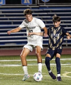 Two high school soccer players compete for the ball on a well-lit field during an evening match.