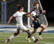 Two high school soccer players compete for the ball during a nighttime match, showcasing athleticism on the field.