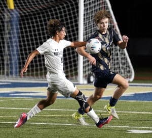 Two high school soccer players compete for the ball during a nighttime match, showcasing athleticism on the field.