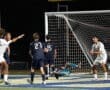 Players celebrate a goal during a nighttime soccer match, with the goalie on the ground and teammates celebrating the score.
