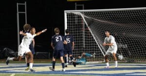 Players celebrate a goal during a nighttime soccer match, with the goalie on the ground and teammates celebrating the score.