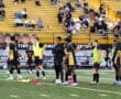 Youth soccer team warming up on the field, dressed in black and gold uniforms, with coaches and spectators in background.