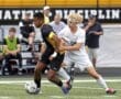 High school soccer players compete for the ball during an intense match on a field. Fans watch in the background.