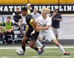High school soccer players compete for the ball during an intense match on a field. Fans watch in the background.