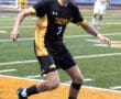 High school soccer player in black and gold uniform dribbles a soccer ball on the field during a match.