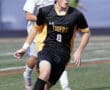 High school soccer player in black jersey dribbles the ball while a teammate pursues from behind on the field.