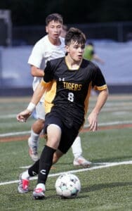 High school soccer player in black jersey dribbles the ball while a teammate pursues from behind on the field.