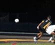 Soccer players in an intense match; one kicks the ball towards the goal while the opponent reacts. Nighttime setting.