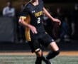 High school soccer player in a black and yellow uniform dribbling a ball during a night game on an outdoor field.