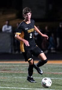 High school soccer player in a black and yellow uniform dribbling a ball during a night game on an outdoor field.