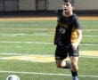 A soccer player in a black and gold uniform sprinting on a field during a night game, focused on the ball ahead.