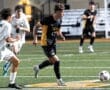 A high school soccer player in a black and yellow uniform dribbles the ball, while opponents pursue him on the field.