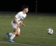 High school soccer player in action, sprinting on the field chasing a ball during a nighttime match.