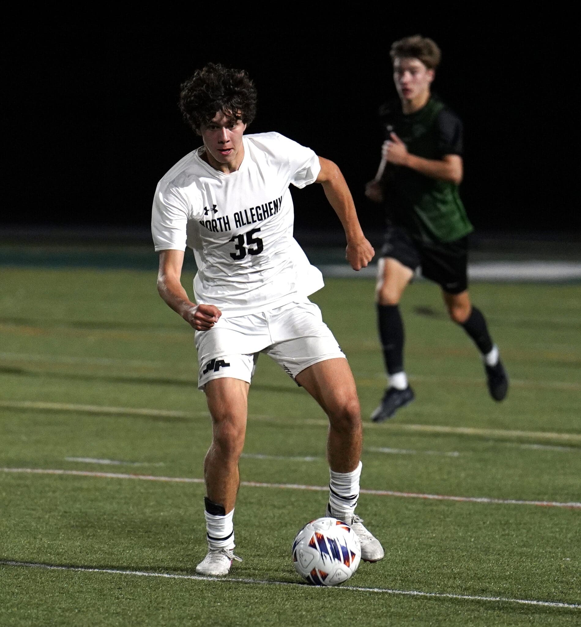 Player in a white jersey with North Allegheny dribbles a soccer ball on a field during a nighttime match.