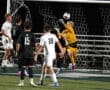 Goalkeeper saves a high shot during a soccer match, with players in black and white uniforms visible on the field.