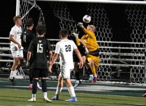 Goalkeeper saves a high shot during a soccer match, with players in black and white uniforms visible on the field.