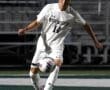 North Allegheny soccer player in white uniform dribbles the ball during a nighttime game on a turf field.