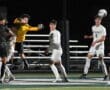 Goalkeeper in yellow leaps to catch the ball during a night soccer match, surrounded by players in white and black uniforms.