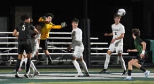 Goalkeeper in yellow leaps to catch the ball during a night soccer match, surrounded by players in white and black uniforms.