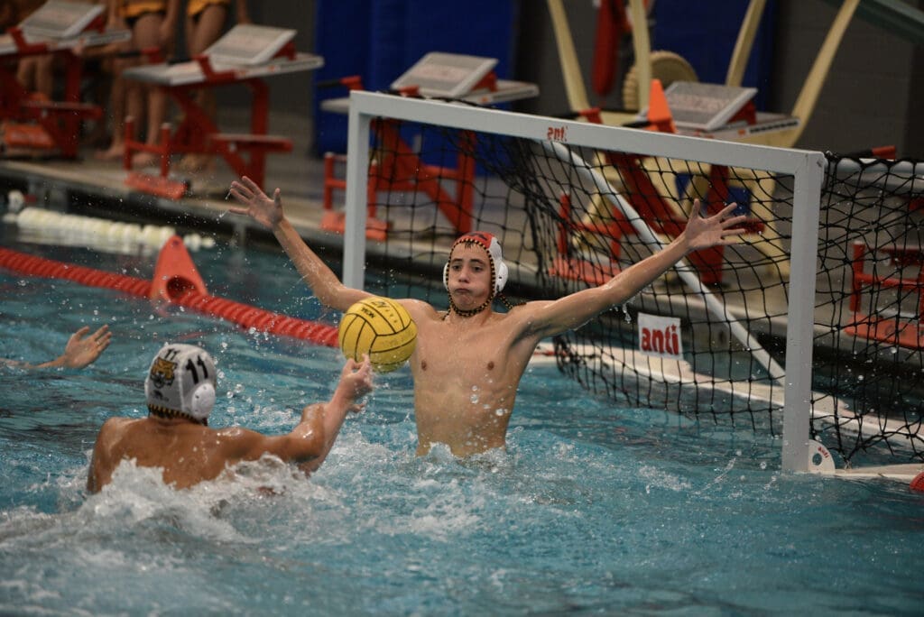 Water polo match in action, with a goalkeeper defending against a shot from an opponent in a splash-filled pool.