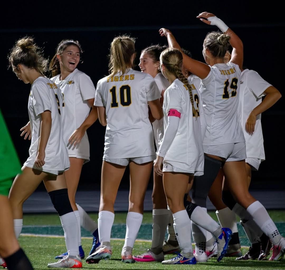 Soccer team celebrating a goal, wearing white jerseys with 'Tigers' on the back, under stadium lights.