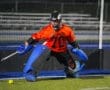 Goalkeeper in orange jersey and blue gear defends the goal during a nighttime field hockey match.