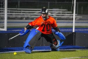 Goalkeeper in orange jersey and blue gear defends the goal during a nighttime field hockey match.