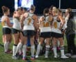 Field hockey team huddling during a night game, focused on strategy and teamwork under the stadium lights.