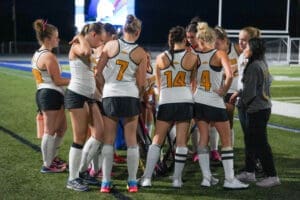 Field hockey team huddling during a night game, focused on strategy and teamwork under the stadium lights.