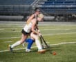 Two field hockey players compete on a turf field, focusing on a red ball while showcasing athletic skill and teamwork.