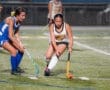 Two young athletes compete in a field hockey match, focused on controlling the ball on a turf field under stadium lights.
