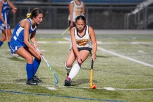 Two young athletes compete in a field hockey match, focused on controlling the ball on a turf field under stadium lights.