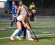 Field hockey players compete for the ball during a night game on a well-lit turf field.