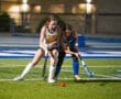 Two female field hockey players compete for the ball on a brightly lit turf field during a game.