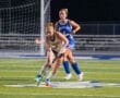 Two high school field hockey players compete for the ball on a brightly lit turf field during a night game.