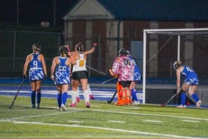 Field hockey players celebrate a goal under stadium lights, with a goalkeeper ready in vibrant gear.