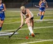 Field hockey players in action; one player stretches to hit the ball while another pursues in blue uniform on turf.