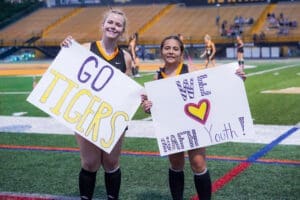 Two young fans cheer at a sports event, holding signs that say Go Tigers and We ♥ Math Youth! on the field.