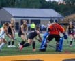 High school field hockey players in action during a game, featuring a goalie in orange and teammates in black uniforms.