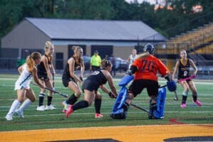 High school field hockey players in action during a game, featuring a goalie in orange and teammates in black uniforms.