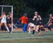 Field hockey players compete for the ball near the goal, with the goalie in orange defending against an approaching attack.