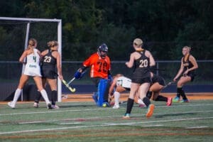 Field hockey players compete for the ball near the goal, with the goalie in orange defending against an approaching attack.