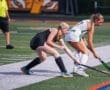 Two female field hockey players compete for the ball on the field during a game, with an official in the background.