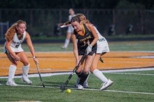 High school girls' field hockey players competing for the ball on the turf during an evening match.