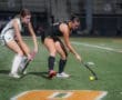 Two female field hockey players compete for the ball on a lit turf field during an evening match.