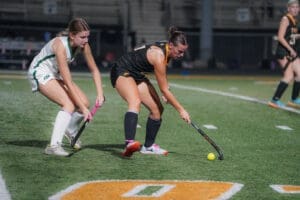 Two female field hockey players compete for the ball on a lit turf field during an evening match.