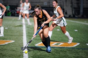 Field hockey players in action, one dribbling a yellow ball while another defender pursues during a night game.