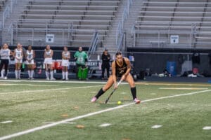 Player in a black jersey prepares to hit a ball during a field hockey match, with teammates in the background.