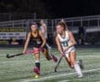 Two high school girls compete in a field hockey match, focusing on the ball under stadium lights at night.