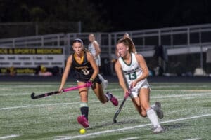Two high school girls compete in a field hockey match, focusing on the ball under stadium lights at night.
